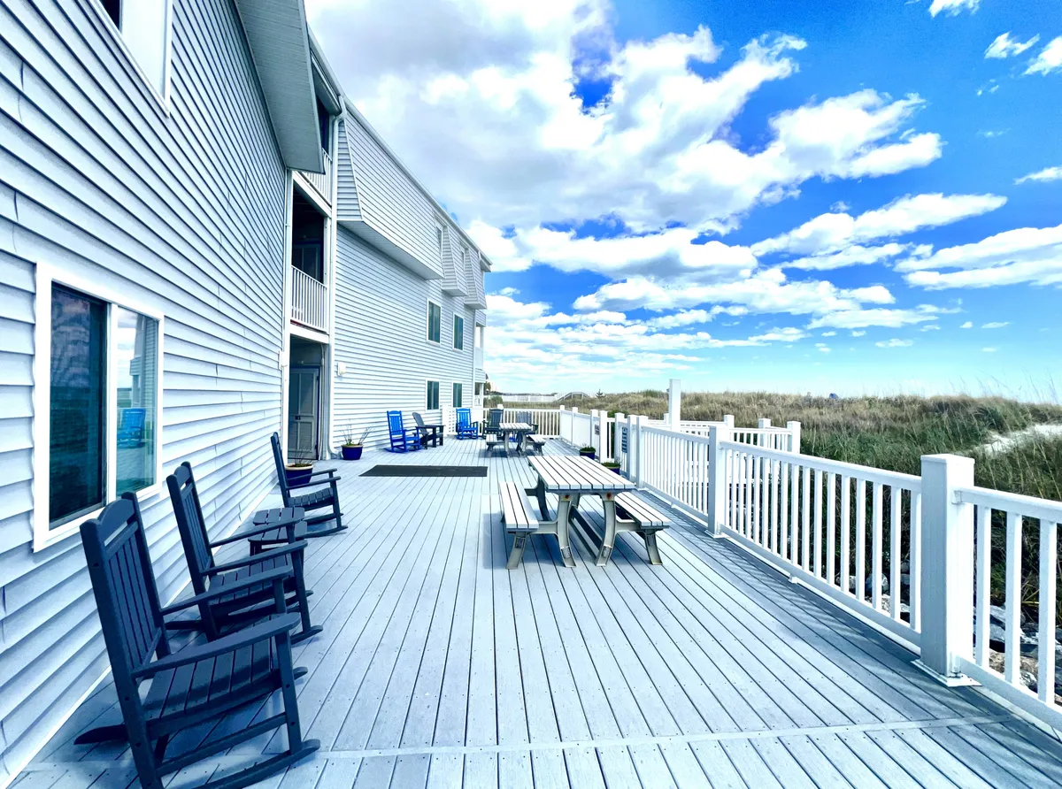 A cheerful exterior scene featuring deck chairs and a picnic table on a spacious outdoor deck, with the building facade and open sky in the background. The scene invites relaxation and socializing, suggesting opportunities for outdoor dining, enjoying morning coffee, or watching sunsets.