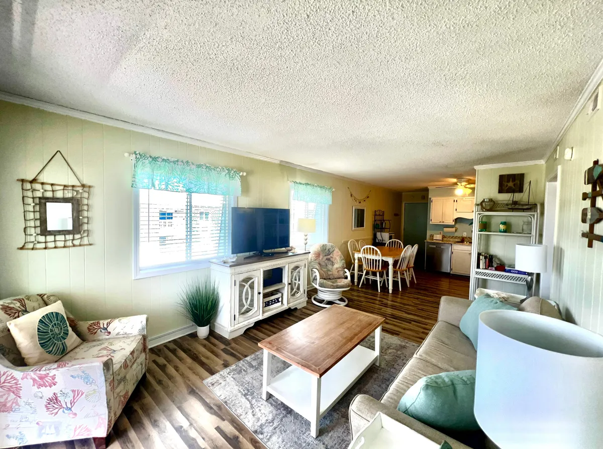 This image captures an open living and dining area with a cozy sofa, coffee table, dining table, and television. The space is filled with natural light, highlighting the plush furnishings and inviting atmosphere. The layout encourages shared meals and relaxation, with décor elements that add elegance.