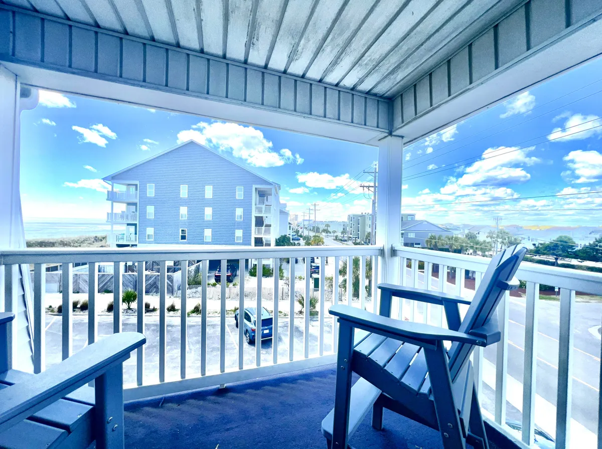 A serene view from the balcony railing, highlighting the proximity to the ocean, a stretch of sand, and the adjacent buildings. The mood is welcoming and calm, suggesting activities like enjoying coffee, reading, or gathering for drinks while taking in the coastal panorama.
