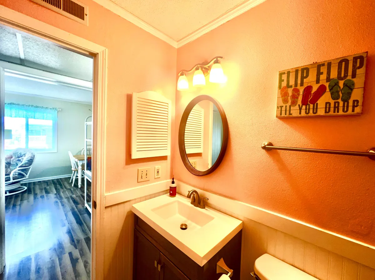 A bright, inviting bathroom with a sink, mirror, modern light fixtures, and a decorative sign, all bathed in morning light. The space feels playful and relaxed, suggesting a spa-like atmosphere ideal for both morning routines and evening unwinding.