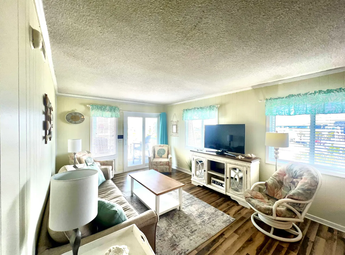 A cozy living area with a comfortable sofa, coffee table, armchair, television, and large windows letting in natural light. The setup evokes a sense of comfort and homelike relaxation, ideal for extended stays and social time.