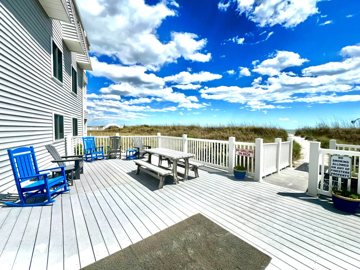 A welcoming deck area featuring blue rocking chairs, a wooden picnic table, and white railings overlooking lush grasses. The space invites relaxation, outdoor meals, and gatherings with friends and family, set against a tranquil coastal backdrop.