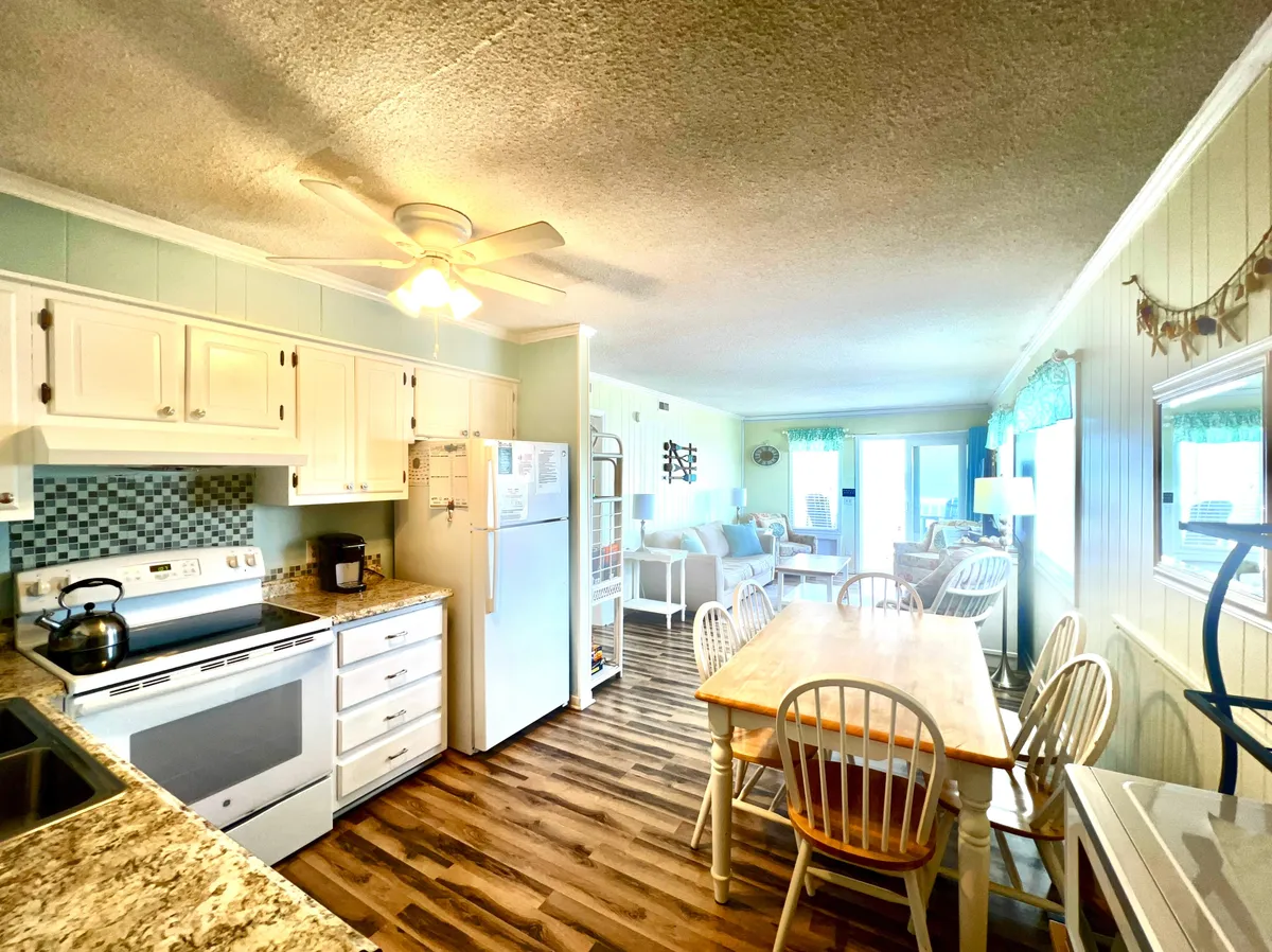 This interior image features the kitchen area with modern appliances, a dining table, and a comfortable seating area. Visible are stainless steel appliances, gleaming countertops, and an airy, open layout that allows for easy movement and interaction. Large windows in the background hint at views beyond, adding a sense of openness and light to the culinary space.