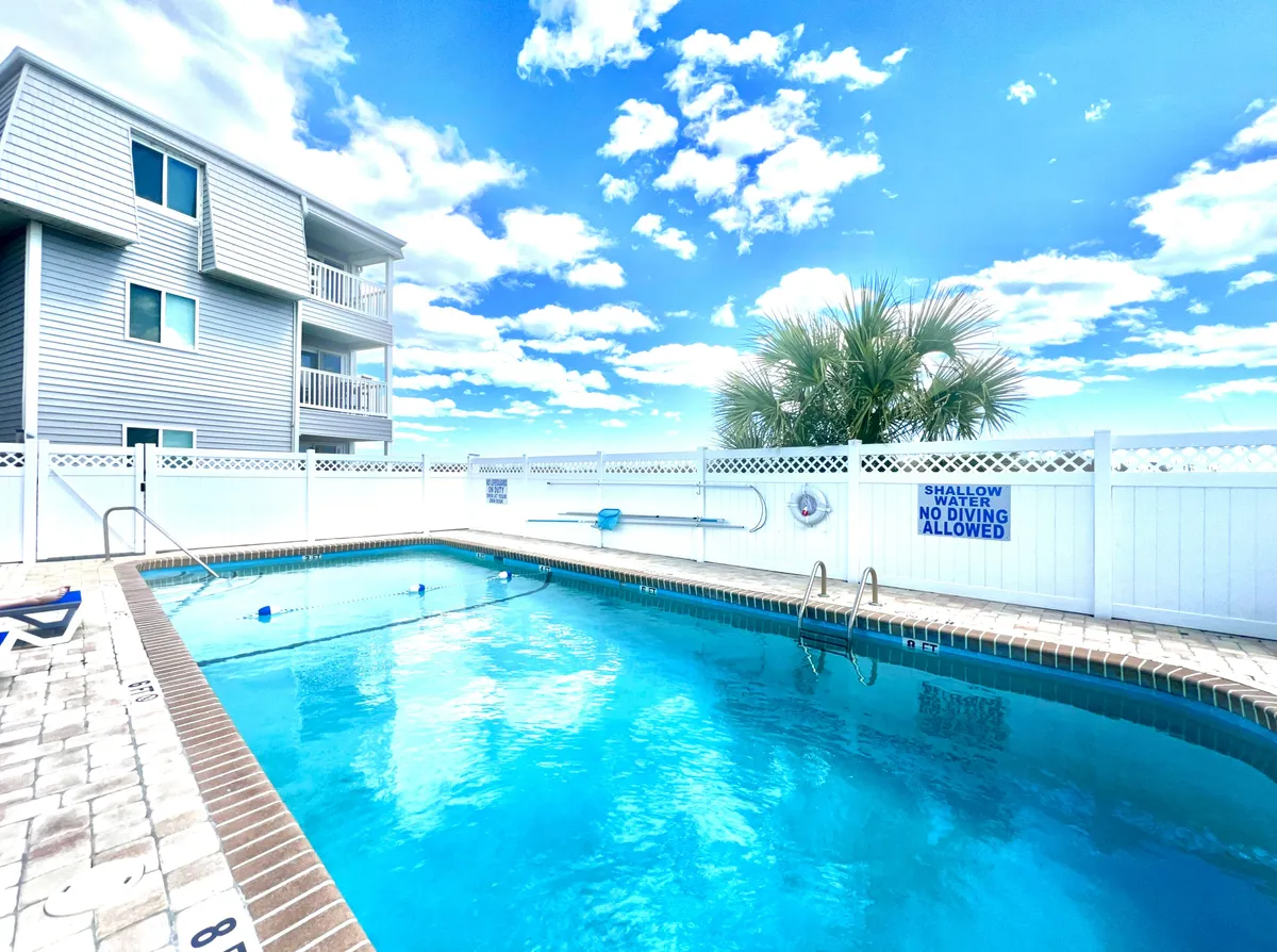 An inviting swimming pool nestled beside the building, with the building’s balconies visible above, palm trees for a resort feel, and plenty of sunlight. The fenced area ensures privacy and safety while the pool area looks perfect for both swimming and lounging.
