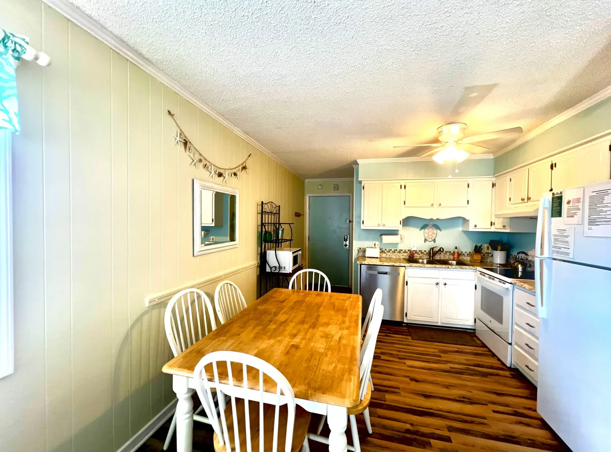 The image features a bright and inviting open-concept living space, centered around a stylish wooden dining table perfect for sharing meals or playing games. White cabinetry and modern kitchen appliances line the background, with a decorative mirror adding a touch of coastal elegance and amplifying the natural light that fills the room. The space radiates warmth and is clearly designed for gathering, relaxation, and family connection.