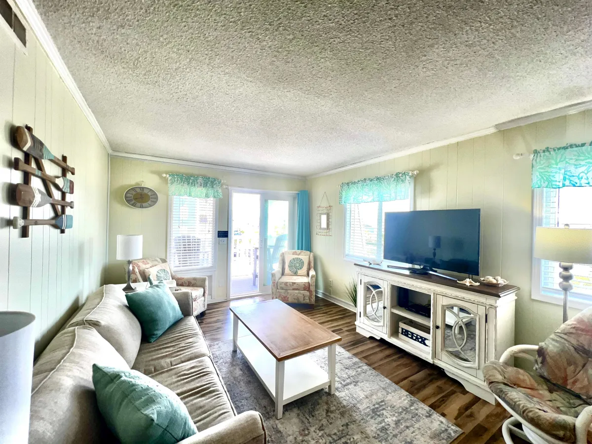 A comfortable living area featuring a plush sofa, coffee table, decorative items, and expansive windows that allow in sunlight. The inviting setup is ideal for relaxing, entertaining guests, or unwinding with a view framed by the windows.