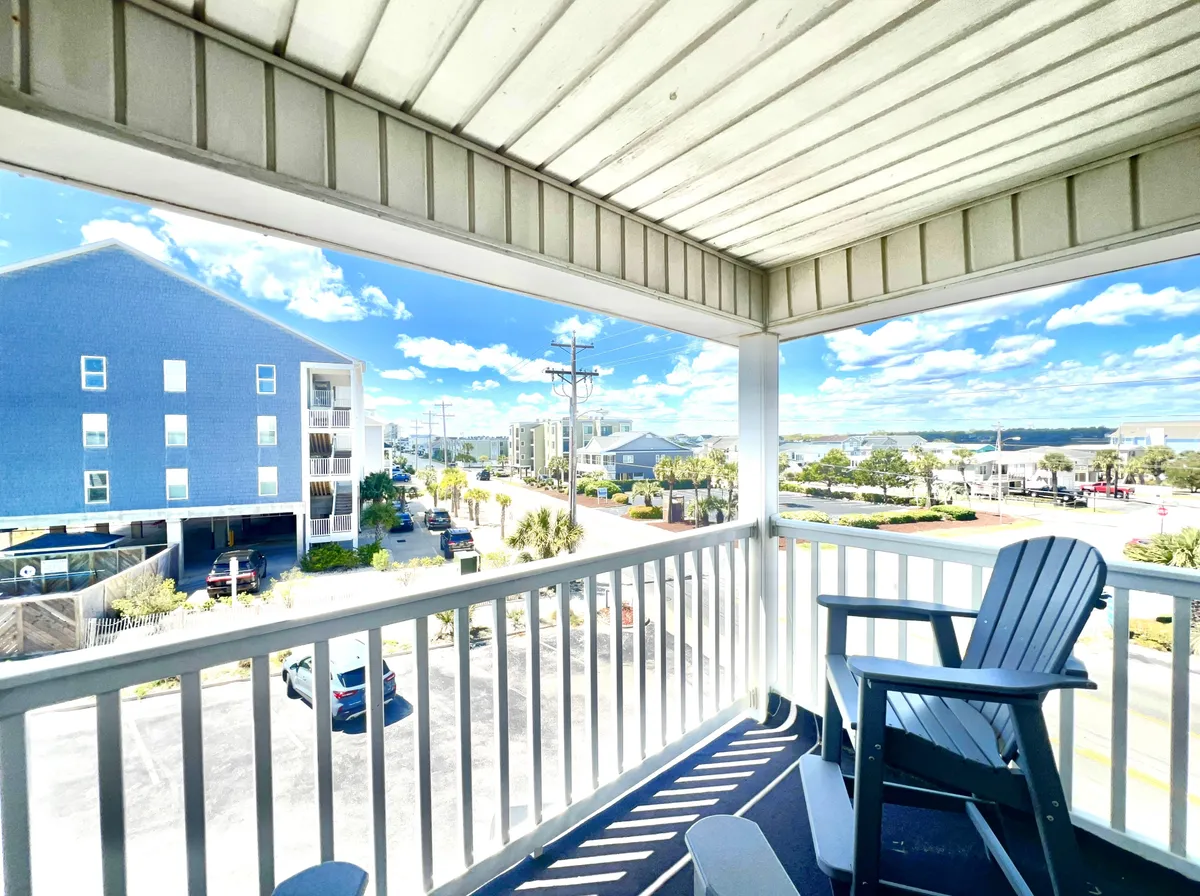 A private balcony featuring an Adirondack chair and railing, overlooking distant buildings and a vast sky. The inviting seating hints at tranquil mornings with coffee or evenings spent watching the sunset together while enjoying ocean breezes.