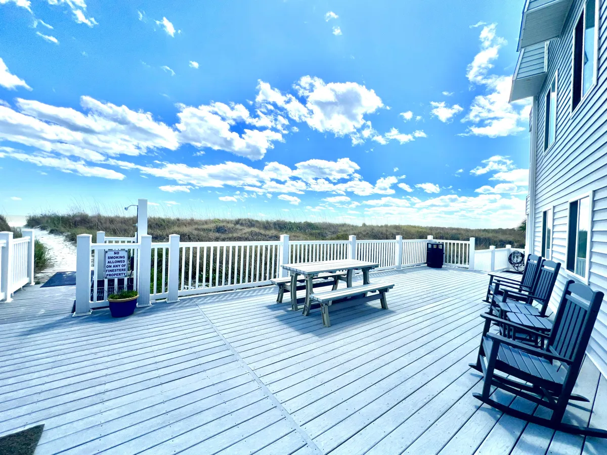 A peaceful outdoor deck with a picnic table and rocking chairs, looking out toward the ocean under a bright sky. The scene suggests opportunities for al fresco dining, sunset drinks, or simply enjoying the sounds of the waves together.