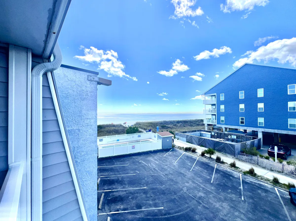 A sweeping ocean view framed by a wide blue sky, with modern buildings below and a parking lot in the foreground. The vantage point suggests proximity to the beachfront, with inviting scenery for beach outings and relaxing moments spent watching the waves.