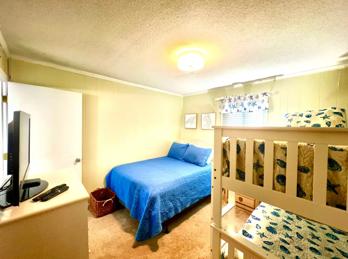 A flexible second bedroom with a blue bedspread, bunk beds, a TV, playful fish-themed wall art, and a window with curtains. The room exudes a relaxed and playful vibe, suitable for kids, guests, or friends, echoing the section's promise of adaptable comfort.